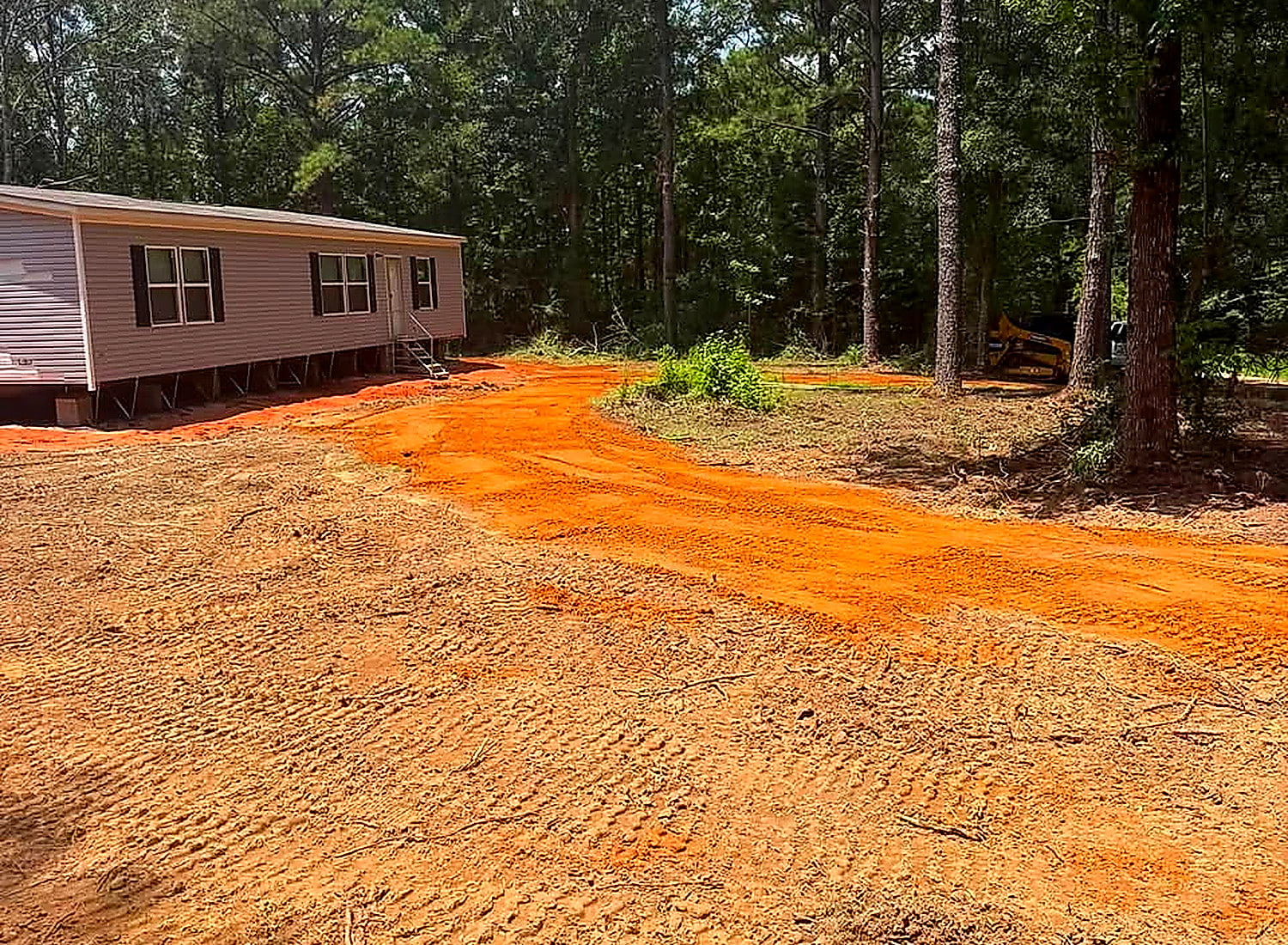 A finished red-dirt pad and driveway in front of a mobile home in the pines