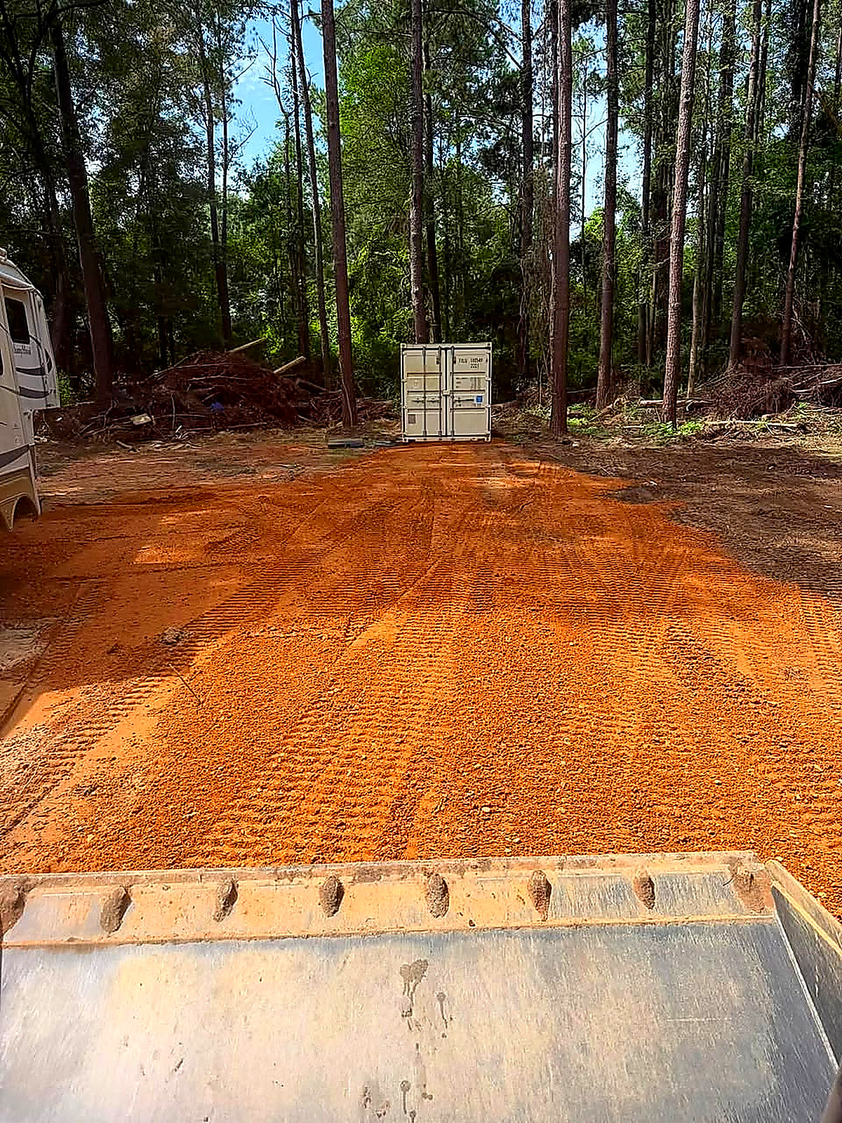 A red-dirt pad with a container set at the back among the pines, seen over the dump trailer tailgate