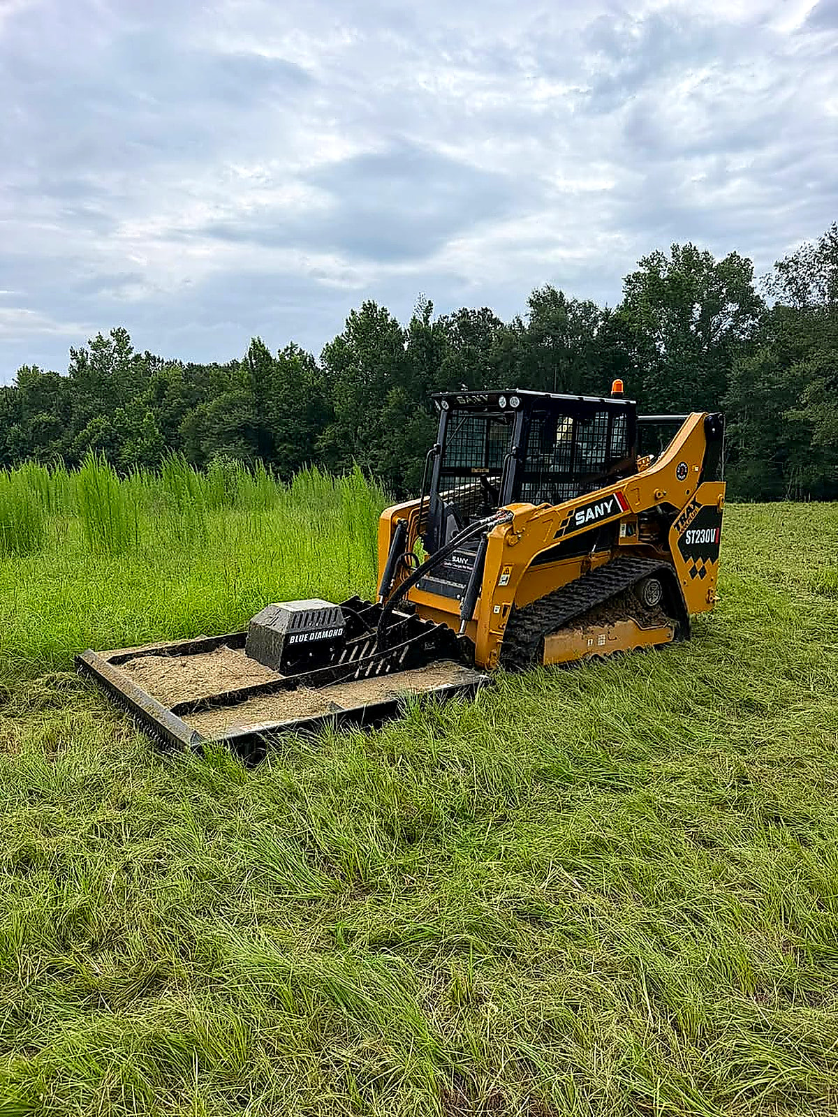 Track loader with a brush cutter working through tall grass on an overcast day