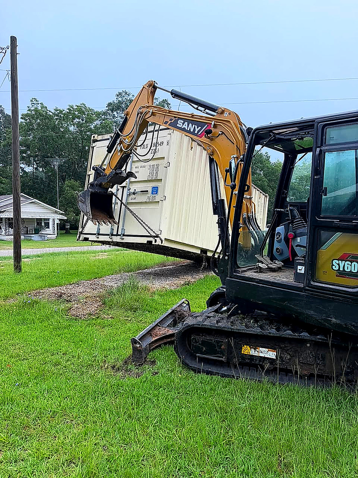A SANY excavator lifting a shipping container beside a house