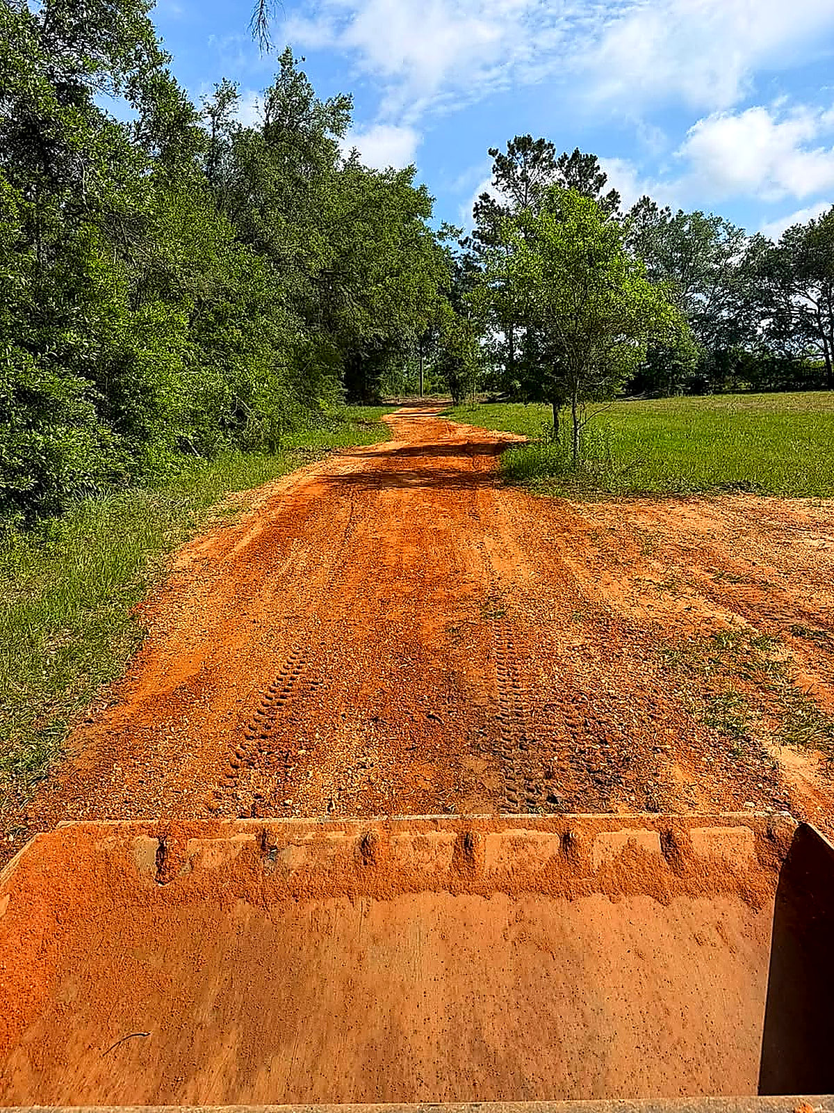 A red-dirt driveway seen out over the loader bucket toward the tree line