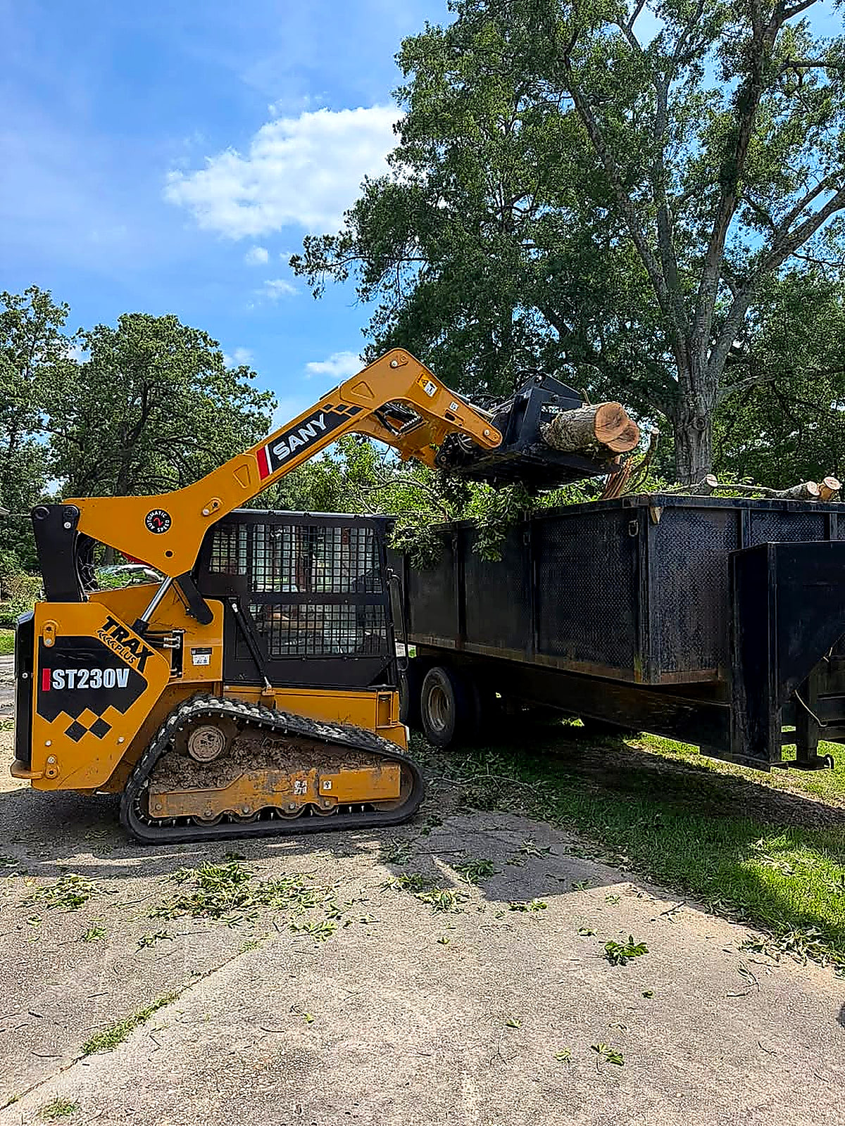 Track loader with a grapple loading storm debris into a dump trailer on a concrete drive
